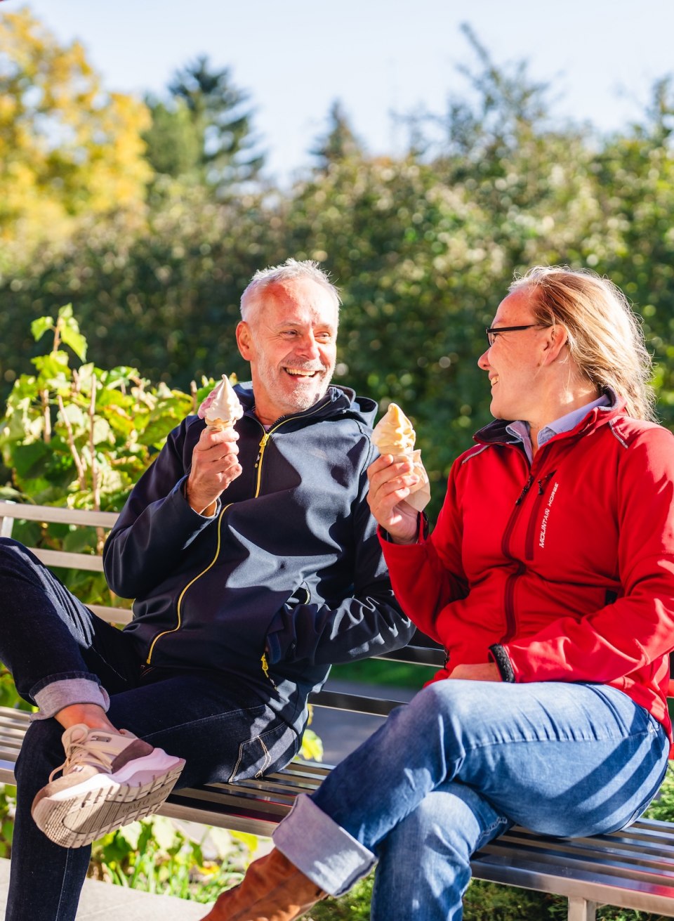 Ein Paar genießt Softeis auf einer Bank in der Sonne, mit grüner Umgebung im Hintergrund.