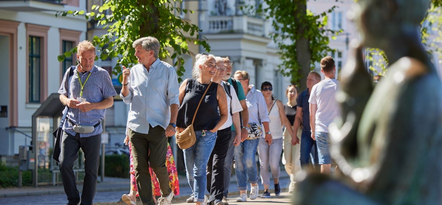 Eine Gruppe Menschen ist mit einem Stadtführer auf einem gepflasterten Weg unterwegs. Rechts, unscharf im Bild ist eine Statue am Pfaffenteich zu sehen, im Hintergrund erscheinen die Villen. Blickpunkt sind der Stadtführer sowie ein Paar, dass sich angeregt unterhält., © Oliver Borchert