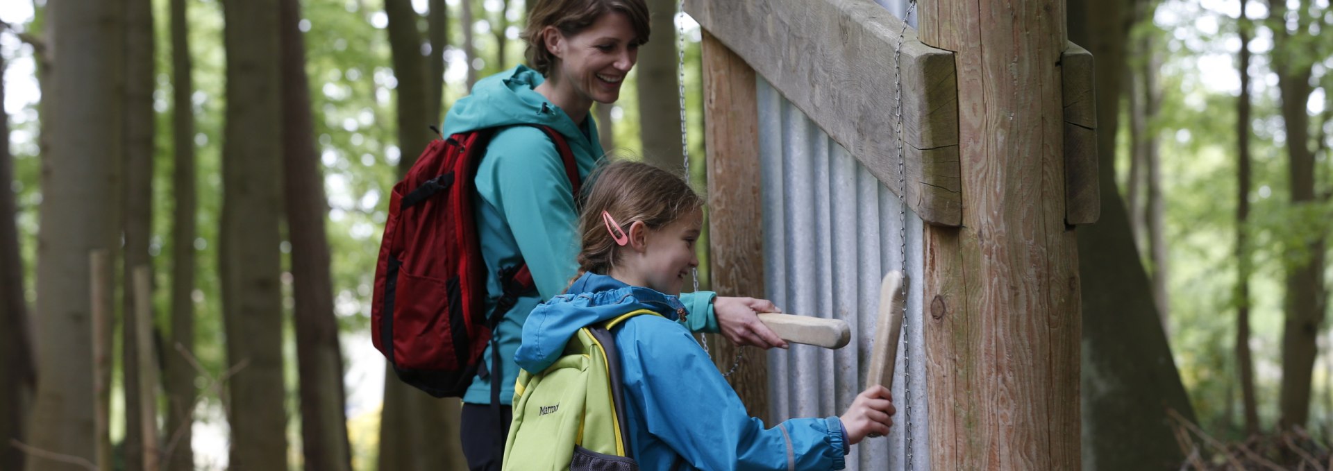 Eine Mutter und ihre Tochter stehen vor einer Installation mit Klangröhren im Klangwald von Graal-Müritz. Beide halten Holzklöppel und schlagen die Röhren an, um Töne zu erzeugen, während sie lächeln.