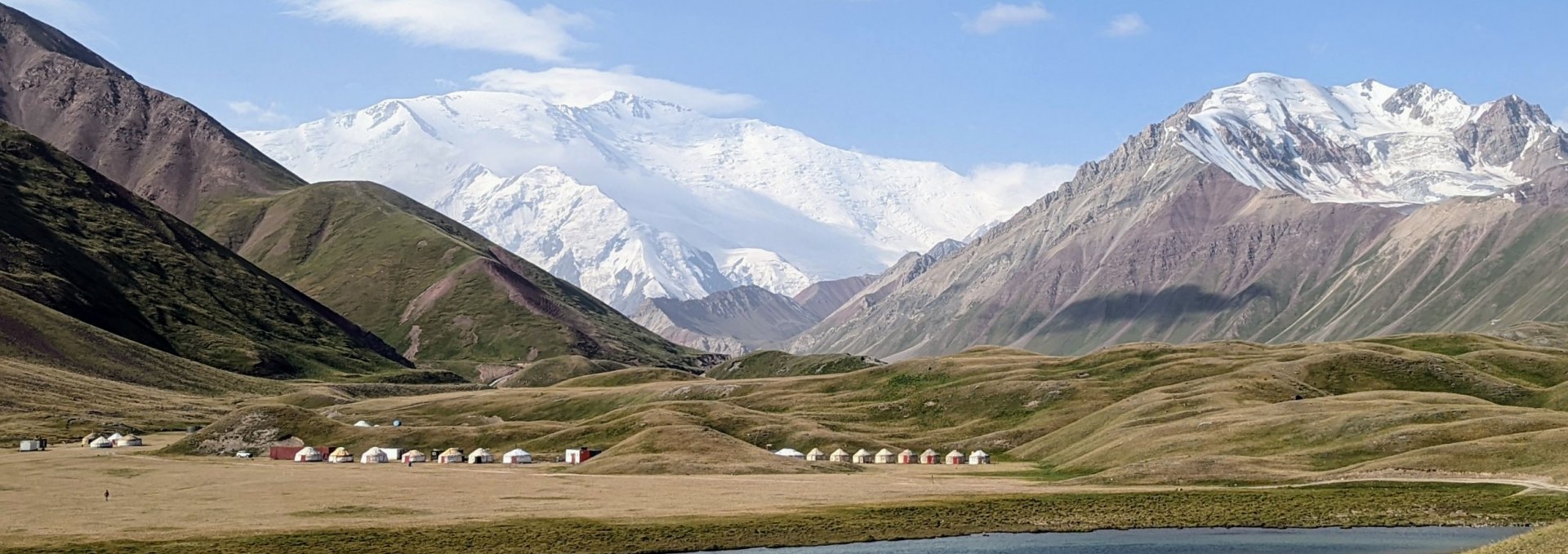 Ein Bild von der Landschaft Kirgistan. Im Hintergrund ist ein Gebirgsmantel, bestehend aus dreu hohen, schneebedeckten Bergen zu sehen.
Davor stehen Hütten von den dortlebenden Menschen und ein Gewässer befindet sich vor ihnen., © Katrin Fischbeck