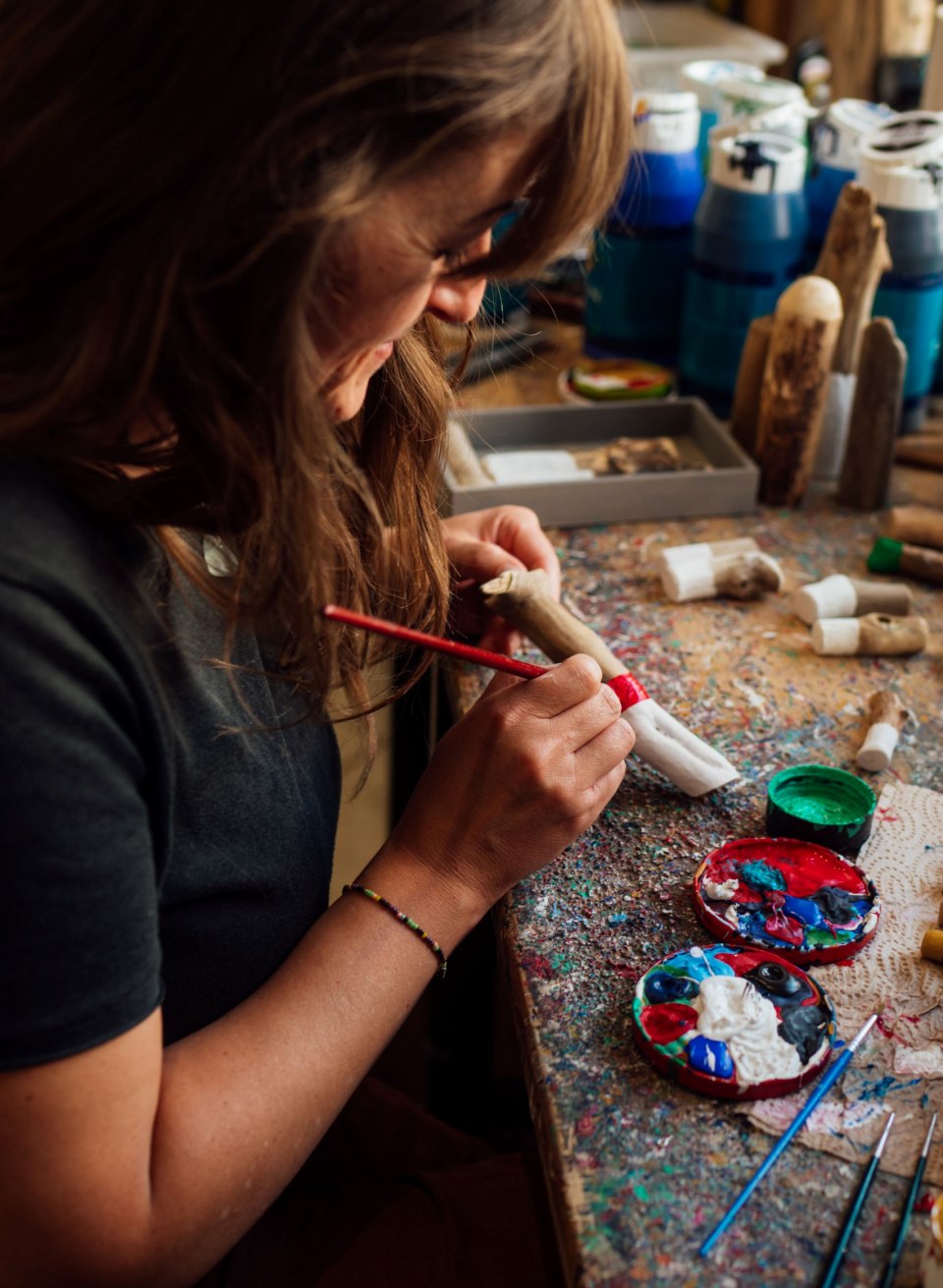 Eine Frau sitzt an einem bunt bemalten Arbeitstisch und bemalt mit einem Pinsel eine Treibholzfigur. Auf dem Tisch liegen Farbtöpfe, Pinsel und weitere kleine Treibholzobjekte, die kunstvoll verziert werden. Im Hintergrund stehen weitere handgefertigte Figuren.