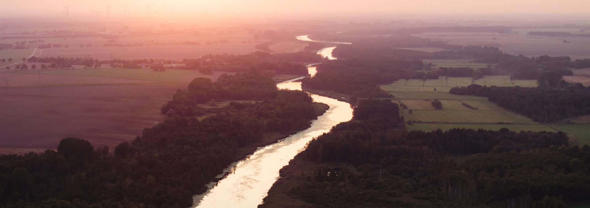 Luftaufnahme der Peene bei Sonnenuntergang, eingebettet in Felder und Wälder, mit einem orangefarbenen Himmel im Hintergrund.