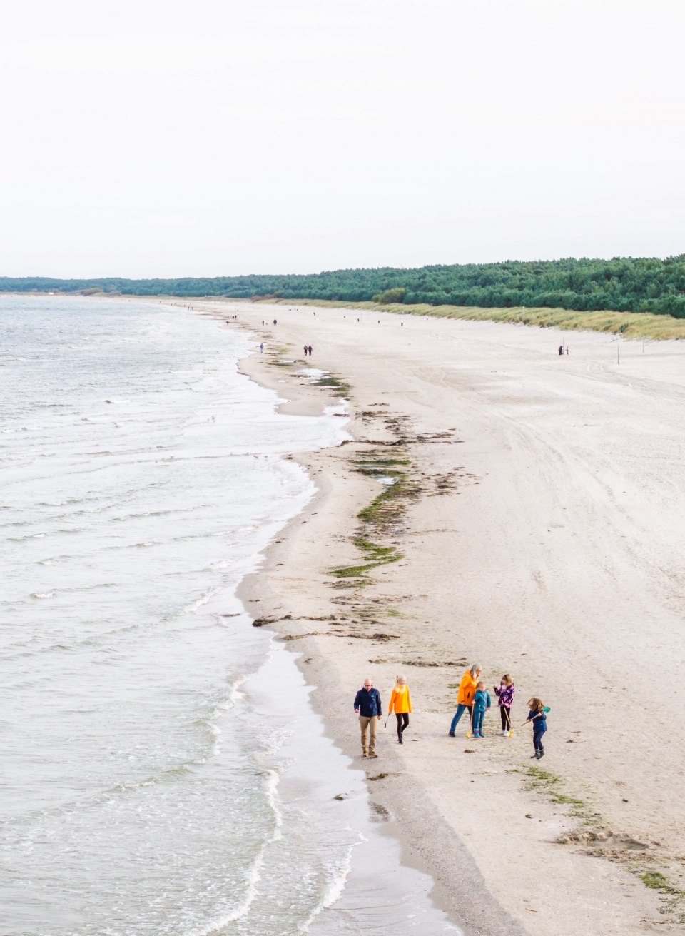 Luftaufnahme eines langen, sandigen Strandes, an dessen Ufer eine Gruppe von Menschen spaziert, umgeben von Meer und grünen Dünen.