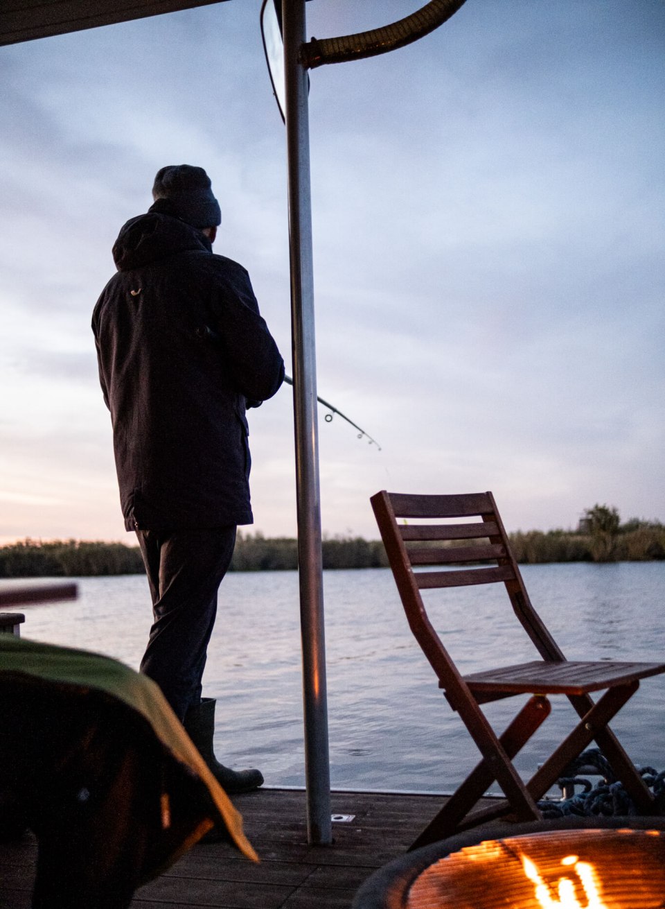 Person angelt vom Deck eines Hausboots auf der Peene bei Sonnenuntergang, mit Feuerstelle im Vordergrund.