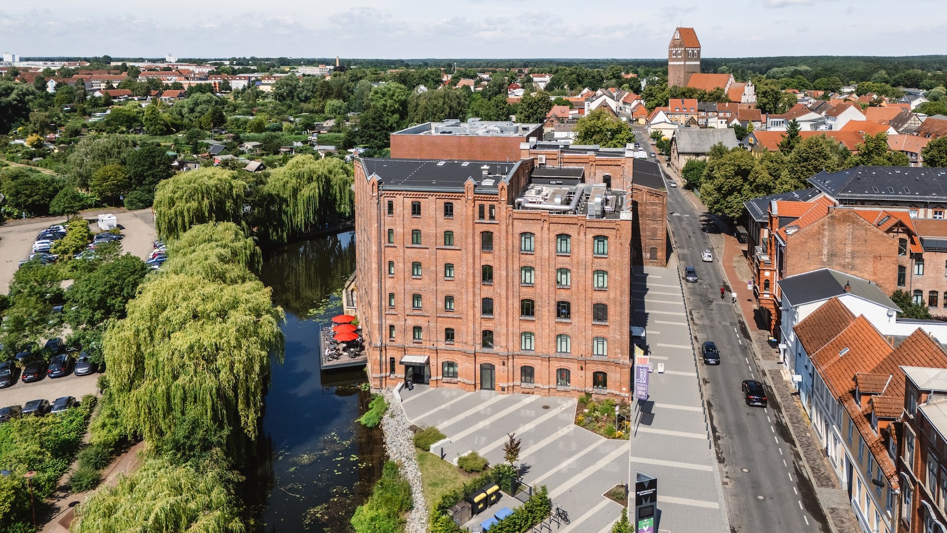 Eine Luftaufnahme zeigt die Kulturmühle in Parchim mit ihrer historischen Fassade und einer gemütlichen Restaurantterrasse am Wasser, umgeben von viel Grün.