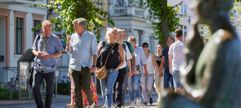 Eine Gruppe Menschen ist mit einem Stadtführer auf einem gepflasterten Weg unterwegs. Rechts, unscharf im Bild ist eine Statue am Pfaffenteich zu sehen, im Hintergrund erscheinen die Villen. Blickpunkt sind der Stadtführer sowie ein Paar, dass sich angeregt unterhält., © Oliver Borchert