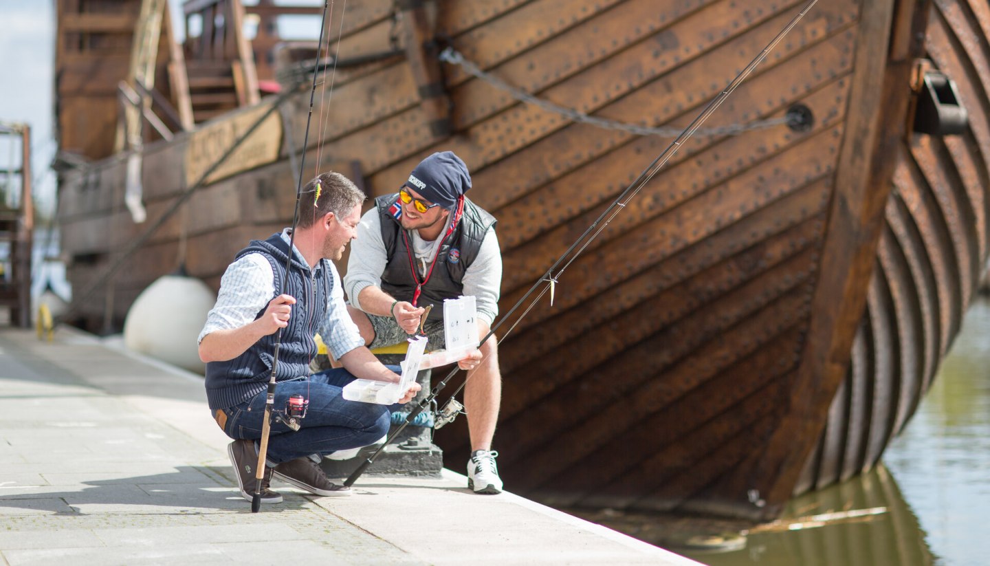 Junge Männer angeln am Stadthafen in Ueckermünde mit einer Kogge im Hintergrund., © TMV/Läufer