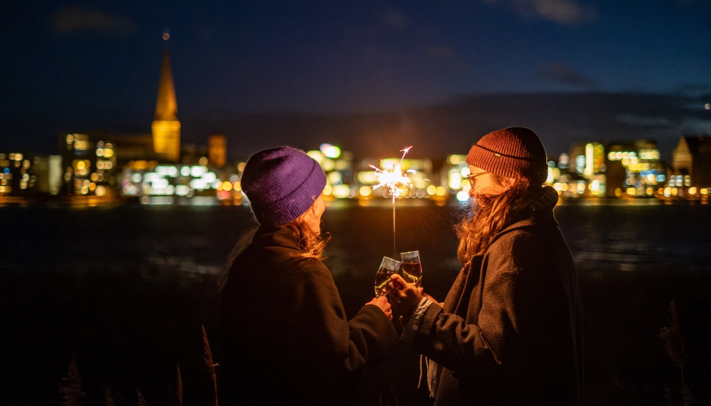 Zwei Personen stoßen mit Sekt an, während sie eine Wunderkerze halten, mit der beleuchteten Skyline von Rostock in Mecklenburg-Vorpommern im Hintergrund.