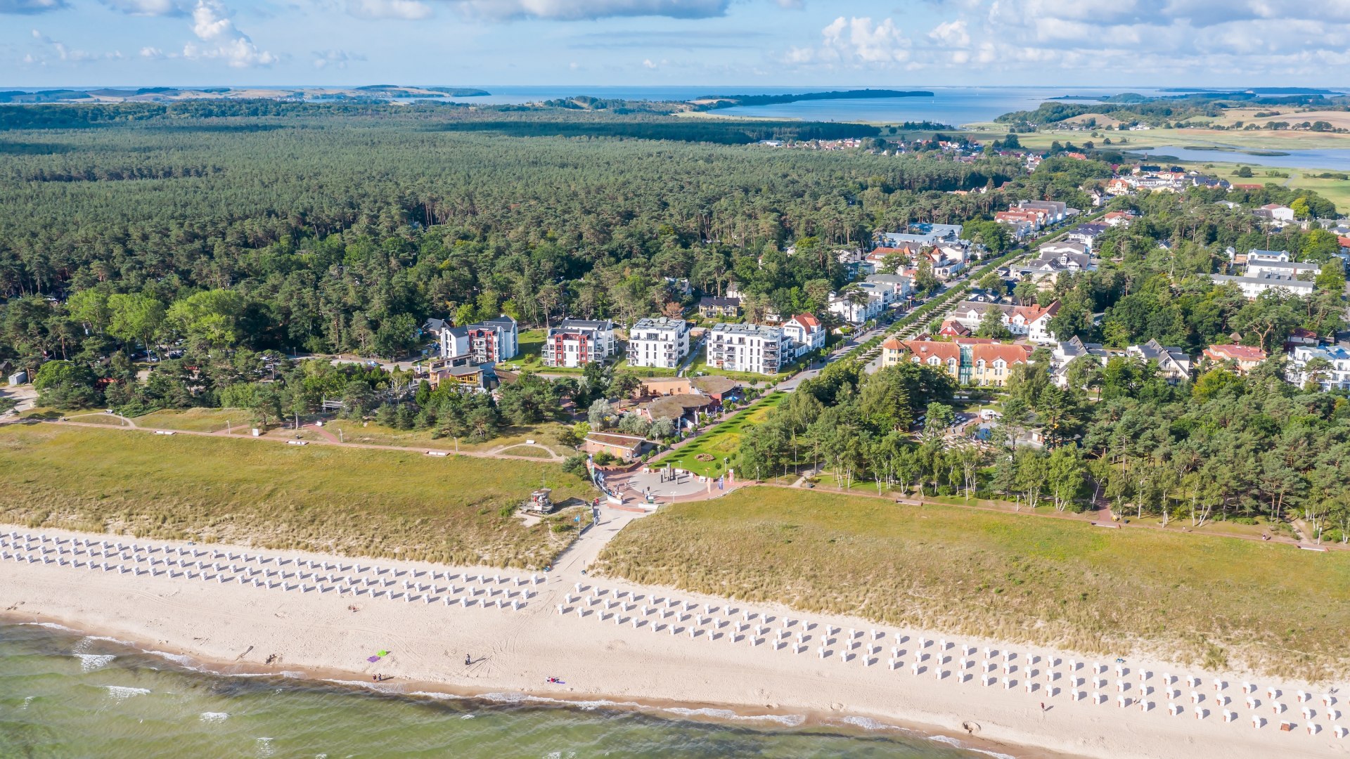 Luftaufnahme des Ostseebads Baabe auf der Insel Rügen: Strand mit Strandkörben, Dünen, moderne Villen und Ferienhäuser, umgeben von dichten Wäldern und Blick auf die Boddenlandschaft.