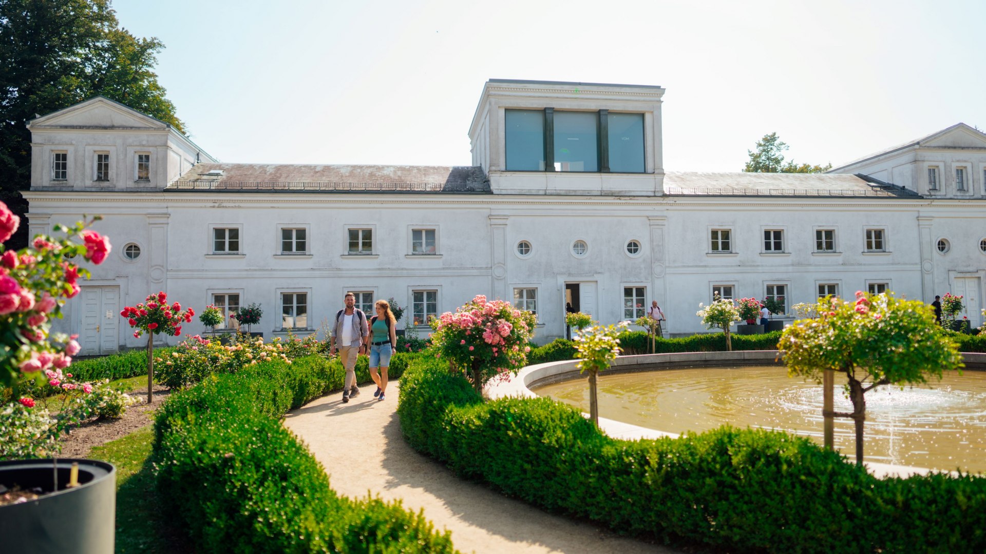 Ein Paar schlendert entlang eines gepflegten Weges durch den Garten der Orangerie in Putbus. Der Garten ist mit Rosen und Buchsbaumhecken geschmückt, während im Vordergrund ein kleiner Teich mit Springbrunnen sichtbar ist. Im Hintergrund steht das historische Gebäude der Orangerie mit weißen Fassaden und großen Fenstern.