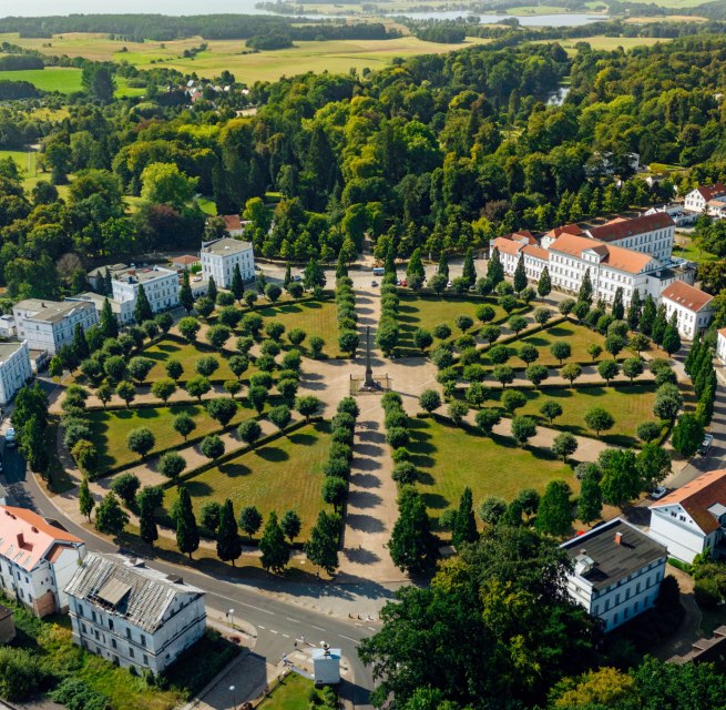 Luftaufnahme des Circus in Putbus auf Rügen, einem kreisförmigen Platz mit symmetrisch angeordneten Bäumen und klassizistischen Gebäuden, umgeben von grüner Natur und ländlicher Landschaft.