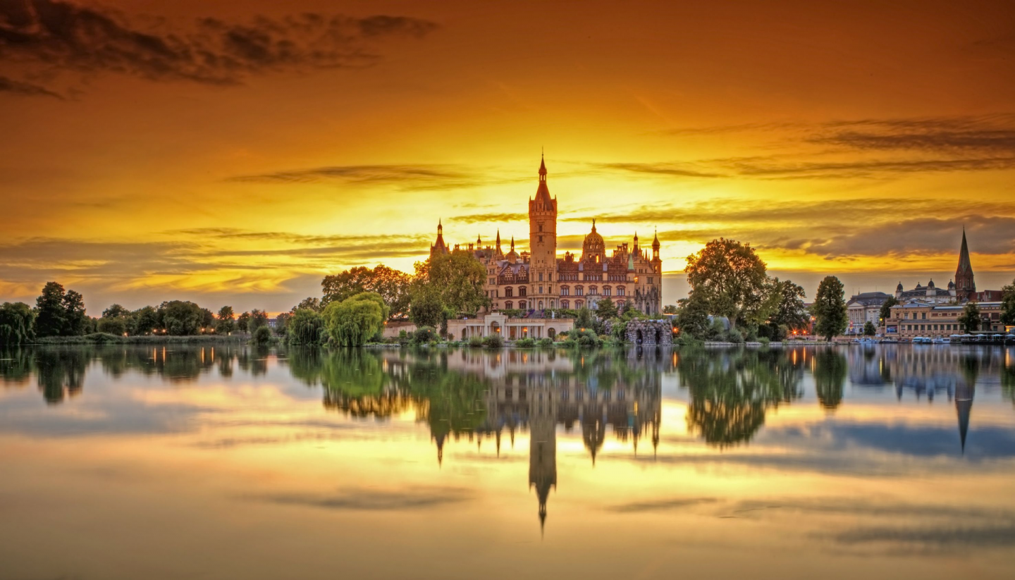 Das Schweriner Schloss wird von den letzten Sonnenstrahlen umrahmt. Der Sonnenuntergang färbt den Himmel orange. Im ruhigen See spiegelt sich das Schweriner Schloss.