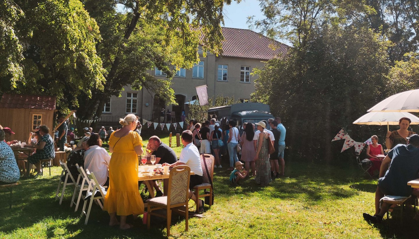 Die MitsommerRemise im Gutspark Klocksin mit Speisen und Getränken. Menschen sitzen an Tischen im Park hinter dem Gutshaus.