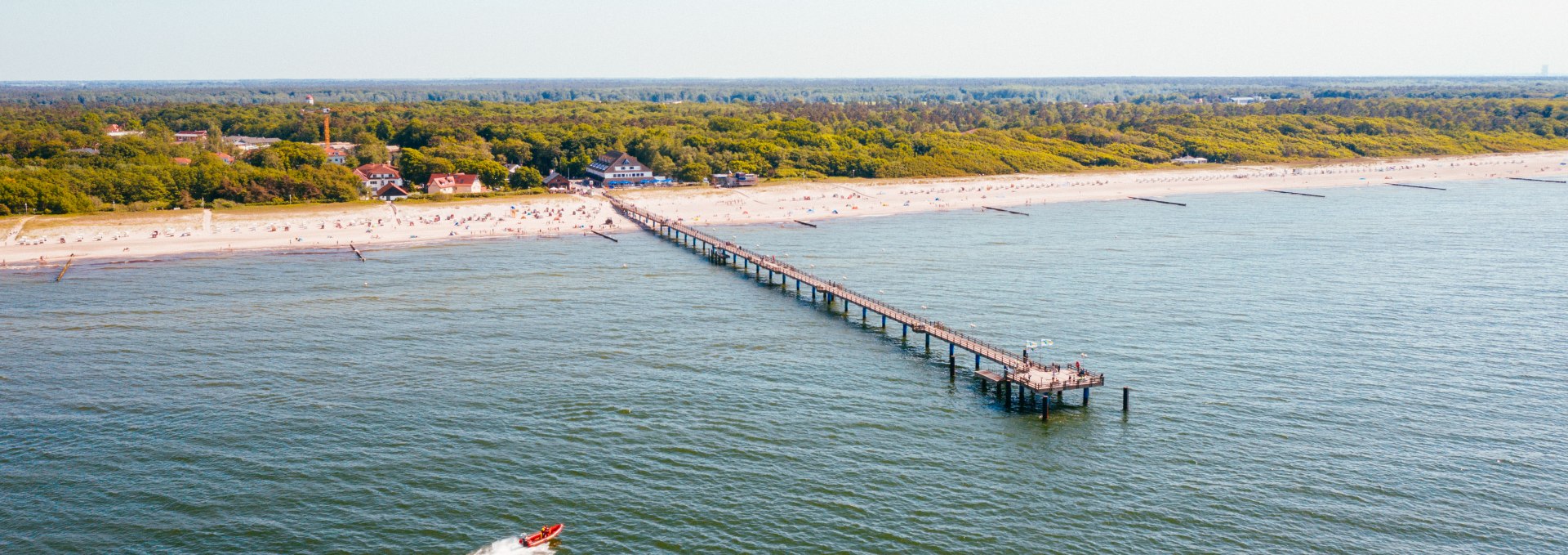 Luftaufnahme der Seebrücke im Ostseeheilbad Graal-Müritz, die weit in die Ostsee hineinragt, umgeben von einem sandigen Strand und dichten grünen Wäldern im Hintergrund.