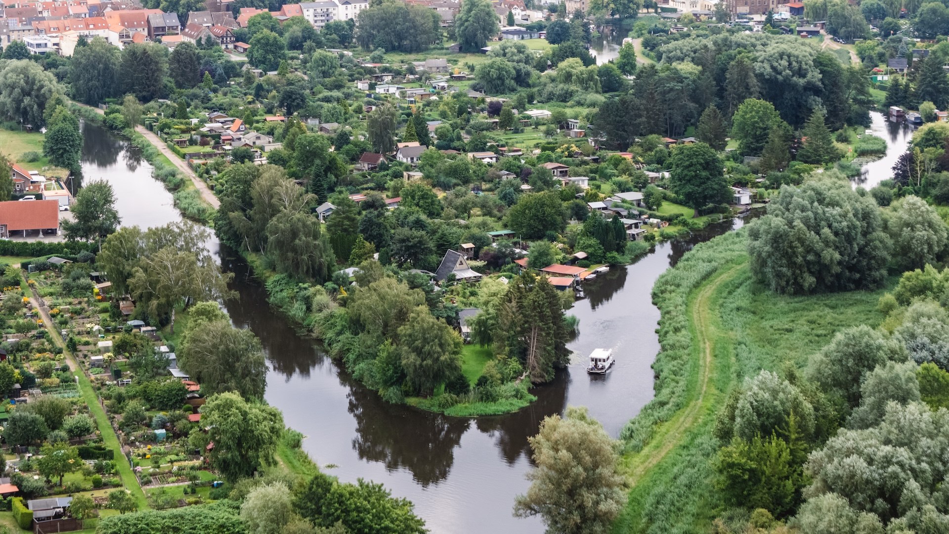 Eine Luftaufnahme zeigt die kurvige Elde mit einem Hausboot, umgeben von grünen Gärten und der Stadt Parchim im Hintergrund.