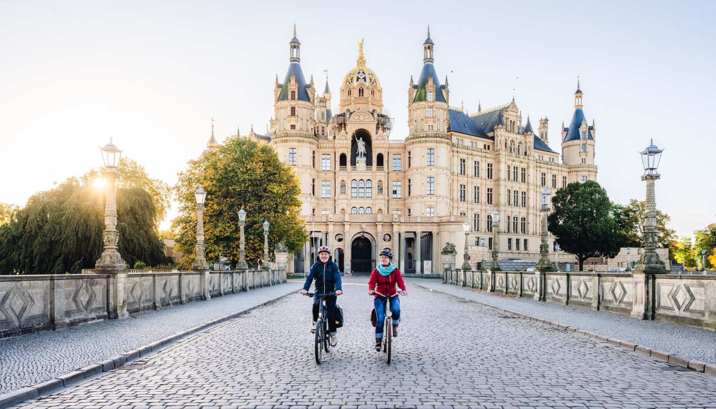 Zwei Radfahrer überqueren eine Brücke vor dem Schweriner Schloss, beleuchtet von der goldenen Morgensonne.