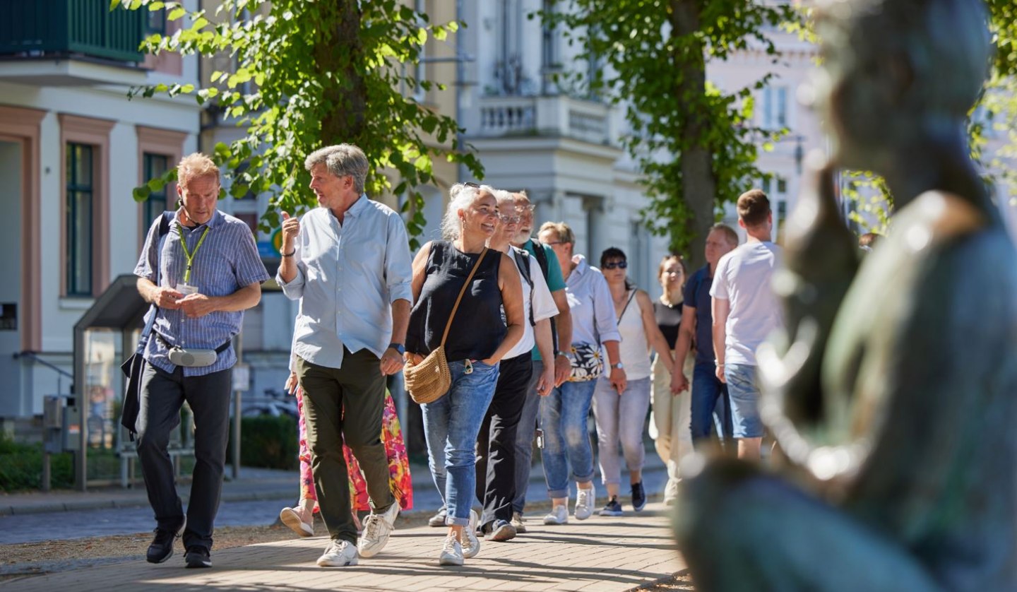 Eine Gruppe Menschen ist mit einem Stadtführer auf einem gepflasterten Weg unterwegs. Rechts, unscharf im Bild ist eine Statue am Pfaffenteich zu sehen, im Hintergrund erscheinen die Villen. Blickpunkt sind der Stadtführer sowie ein Paar, dass sich angeregt unterhält., © Oliver Borchert