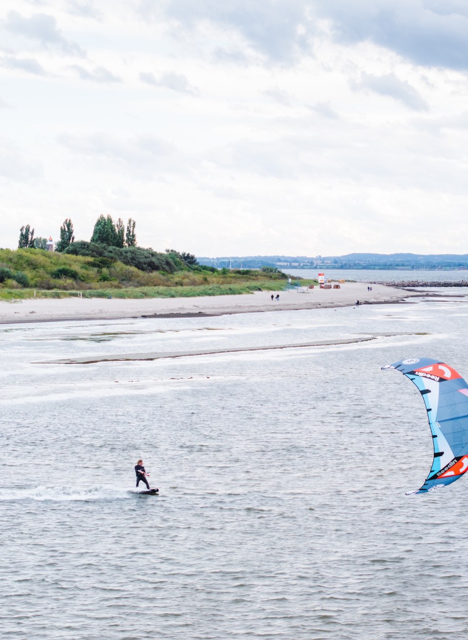 Ein Kitesurfer gleitet vor der Küste von Timmendorf auf Poel über das Wasser, im Hintergrund eine grüne Dünenlandschaft.