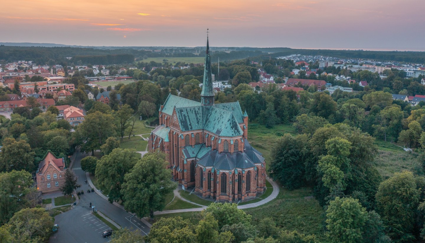 Luftaufnahme des Münsters in Bad Doberan bei Sonnenuntergang, umgeben von Bäumen, Wiesen und der umliegenden Stadt.