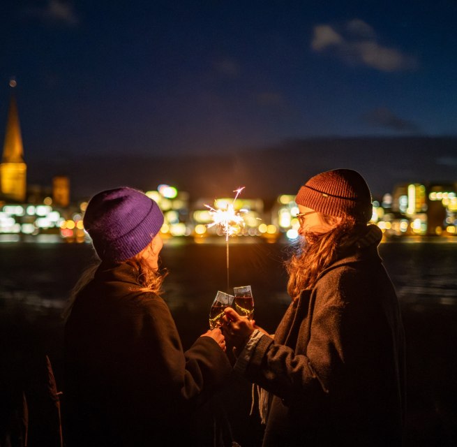 Zwei Personen stoßen mit Sekt an, während sie eine Wunderkerze halten, mit der beleuchteten Skyline von Rostock in Mecklenburg-Vorpommern im Hintergrund.