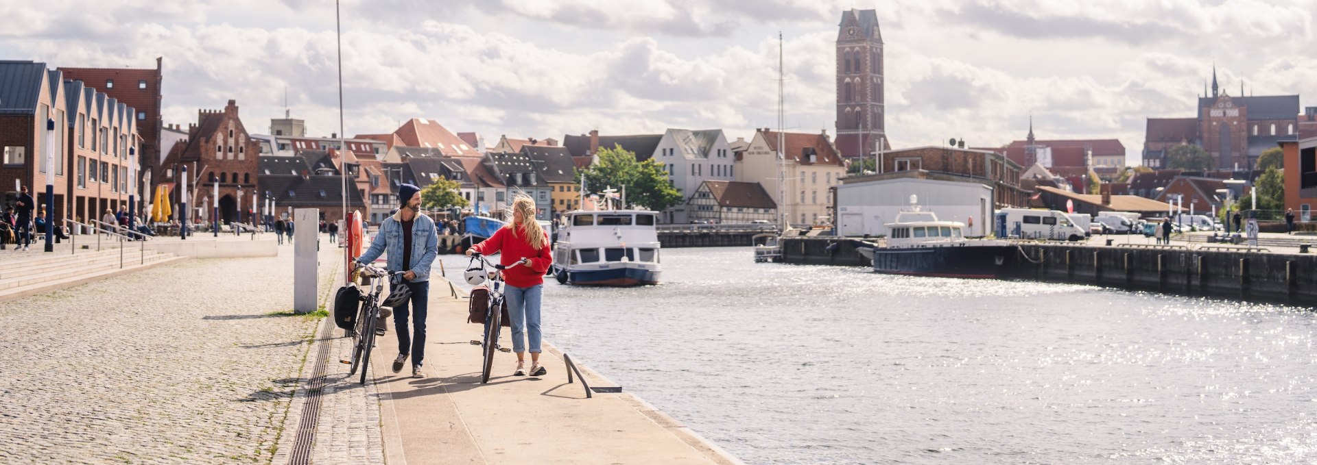 Ein Paar schiebt Fahrräder am alten Hafen von Wismar entlang, im Hintergrund die historische Skyline der Stadt.