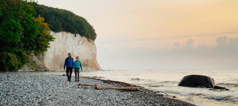 Ein Paar spaziert am Fuße der berühmten Kreidefelsen auf der Insel Rügen entlang, während der Sonnenuntergang den Himmel in sanften Farben erstrahlen lässt.