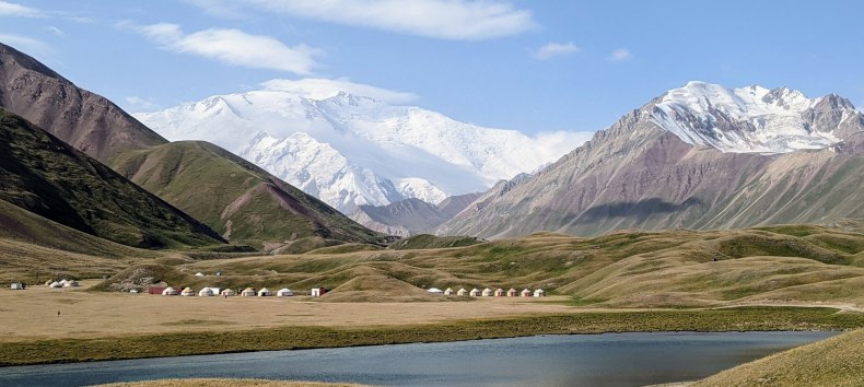 Ein Bild von der Landschaft Kirgistan. Im Hintergrund ist ein Gebirgsmantel, bestehend aus dreu hohen, schneebedeckten Bergen zu sehen.
Davor stehen Hütten von den dortlebenden Menschen und ein Gewässer befindet sich vor ihnen., © Katrin Fischbeck