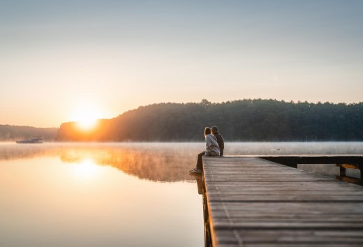 Ein Paar sitzt auf einem Steg zum Sonnenuntergang am Mirower See. Im Hintergrund fährt ein Hausboot vorbei.