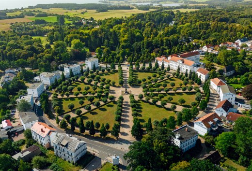 Luftaufnahme des Circus in Putbus auf Rügen, einem kreisförmigen Platz mit symmetrisch angeordneten Bäumen und klassizistischen Gebäuden, umgeben von grüner Natur und ländlicher Landschaft.